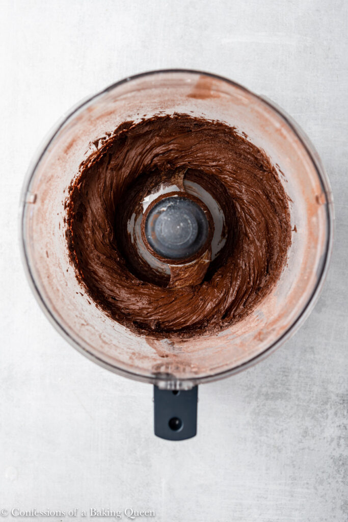 butter, sugar, and cocoa mixed together in a food processor sitting on a grey surface