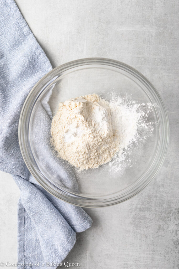 flour, baking powder, baking soda, and salt in a glass bowl on a grey surface with a blue linen