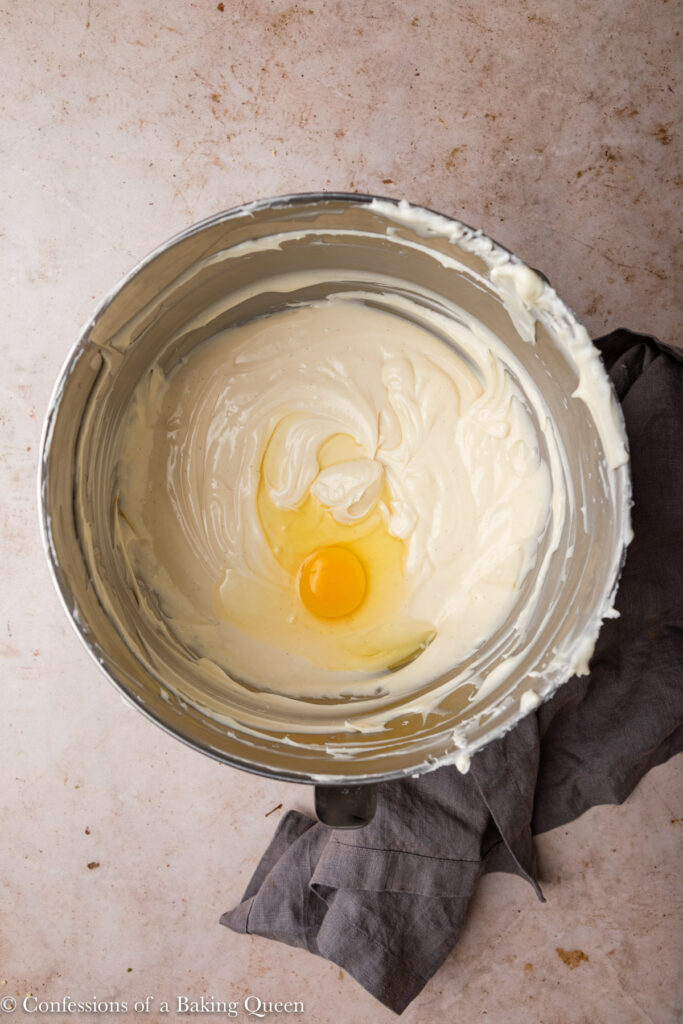 one egg added to cheesecake batter in a metal bowl on a light brown surface with a grey linen