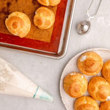 piping bag with whipped cream and a blue piping tip next to a plate full of cream puffs, a baking tray of choux pastry, and a powdered sugar sifter