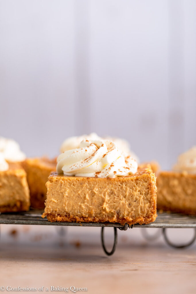 pumpkin cheesecake bars with whipped cream on a wire rack on a brown surface