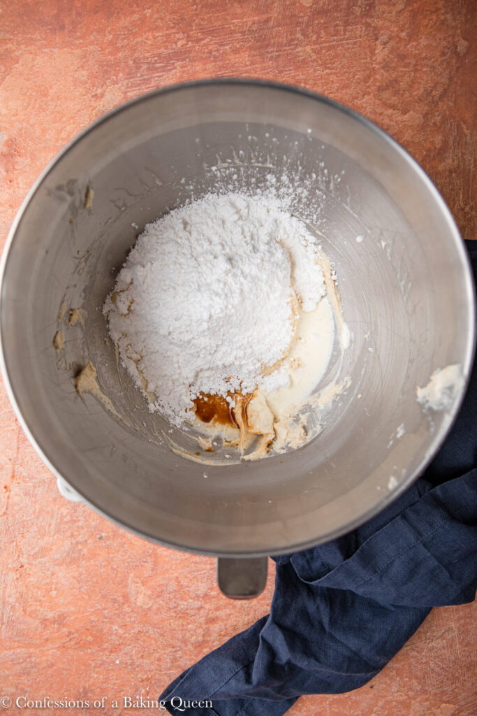 confectioners sugar, vanilla, and heavy cream added to cream cheese butter mixture in a metal mixing bowl on a reddish brown surface with a blue linen