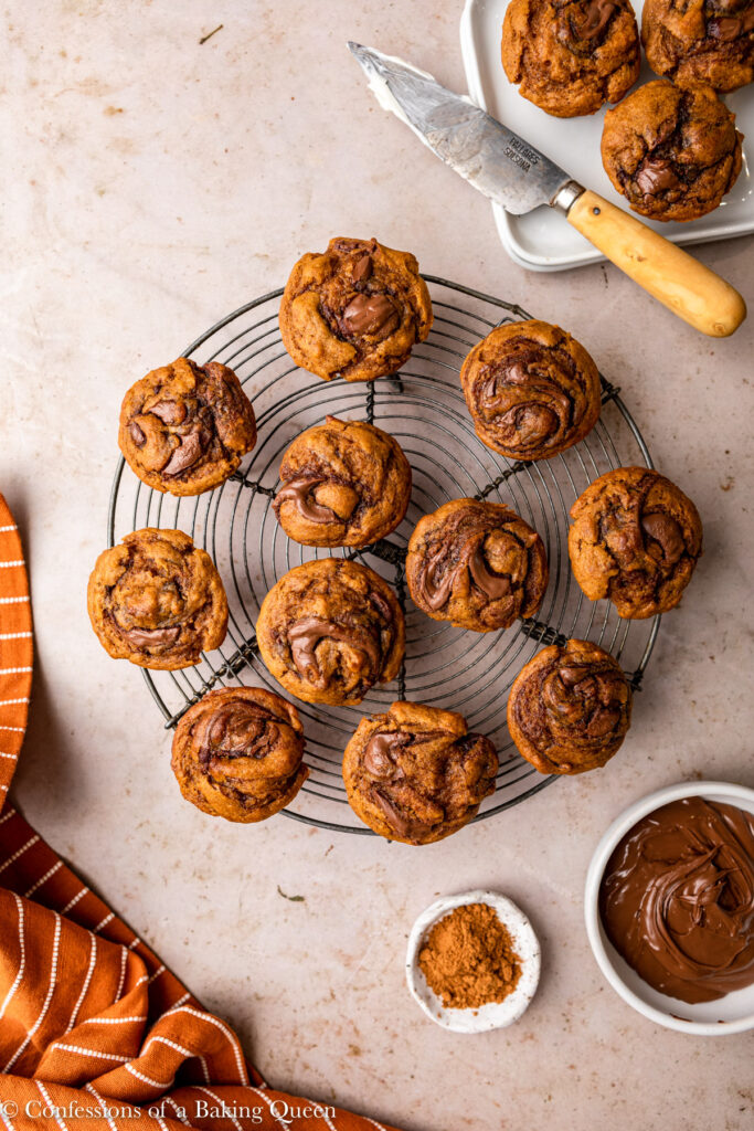 mini pumpkin nutella muffins on a wire rack on a light brown surface with an orange linen, small bowl of nutella, and small plate of pumpkin pie spice