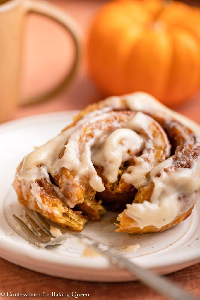 pumpkin cinnamon roll hafl eaten on a white plate with a fork and a cup of coffee and pumpkin in the background on a reddish brown surface