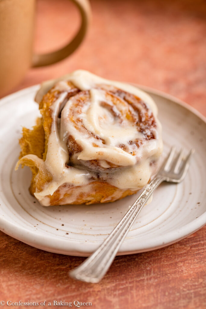 pumpkin cinnamon roll on a white plate with a fork and a cup of coffee in the background on a reddish brown surface