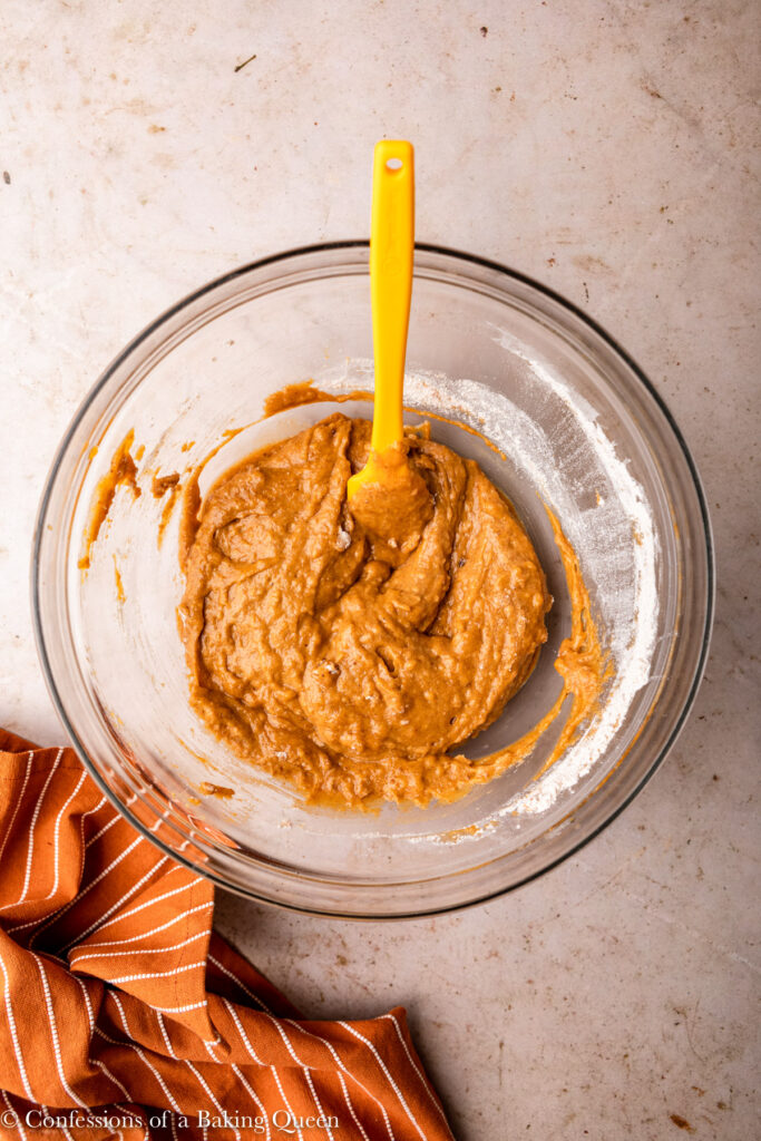 pumpkin muffin batter in a glass bowl with a yellow spatula on a light brown surface with an orange linen