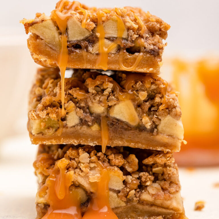 three salted caramel apple pie bars on top of each other with caramel dripping down on a white surface