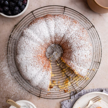 whipping cream bundt cake on a wire rack on a light brown surface with a cup of coffee next to it adn a plate with a fork and slice of cake and a bowl of blueberries