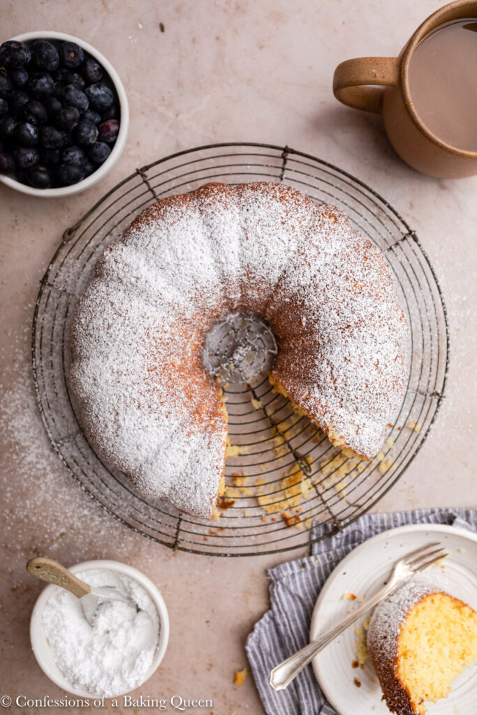 whipping cream bundt cake on a wire rack on a light brown surface with a cup of coffee next to it adn a plate with a fork and slice of cake and a bowl of blueberries 