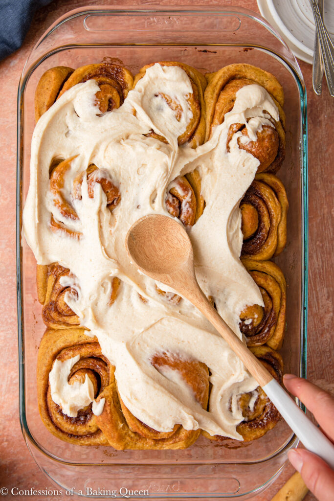 wooden spoon spreading brown butter cream cheese frosting on top of pumpkin cinnamon rolls in a glass dish on a red and brown surface