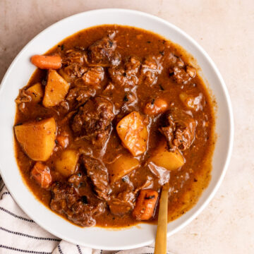 beef stew in a bowl with a spoon next to a white stripped linen on a light brown surface