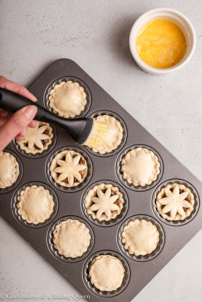 pastry brush spreading egg wash on top of mince pies before baking sitting on a light grey surface with a bowl of egg wash