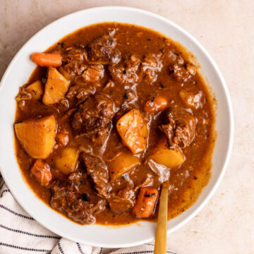 beef stew in a bowl with a spoon next to a white stripped linen on a light brown surface