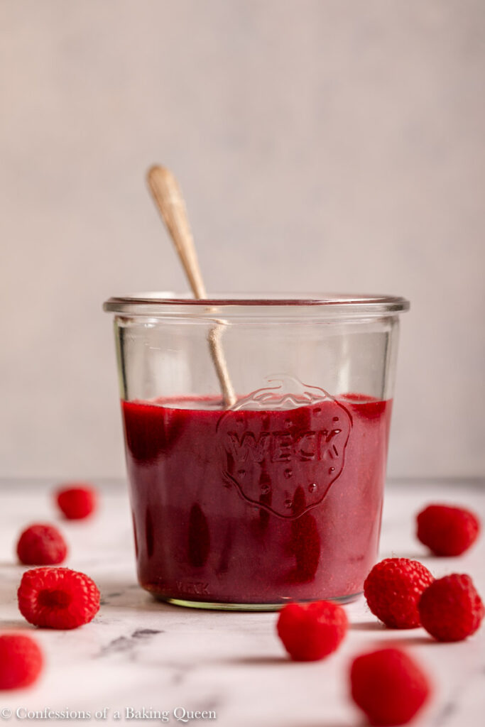 raspberry coulis in a glass jar with a metal spoon on a marble surface with raspberries