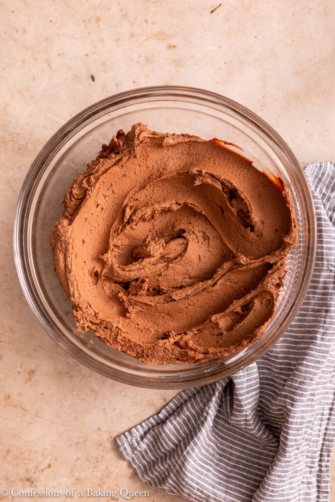 chocolate American buttercream swirled in a glass bowl on a light brown surface with a blue linen