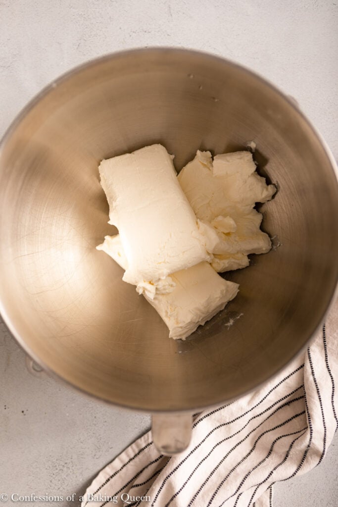 A mixing bowl containing several blocks of cream cheese, ready to be transformed into decadent chocolate cheesecake bars, with a striped kitchen towel partially visible to the right.