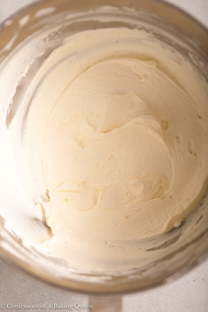 A stainless steel mixing bowl filled with smooth, creamy white frosting—perfect for topping chocolate cheesecake bars—is shown from above.