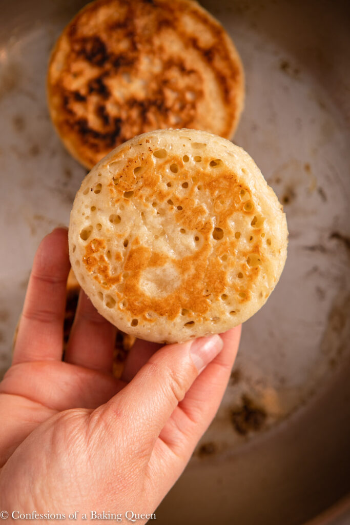 hand holding a crumpet on top of the pan showing its golden brown