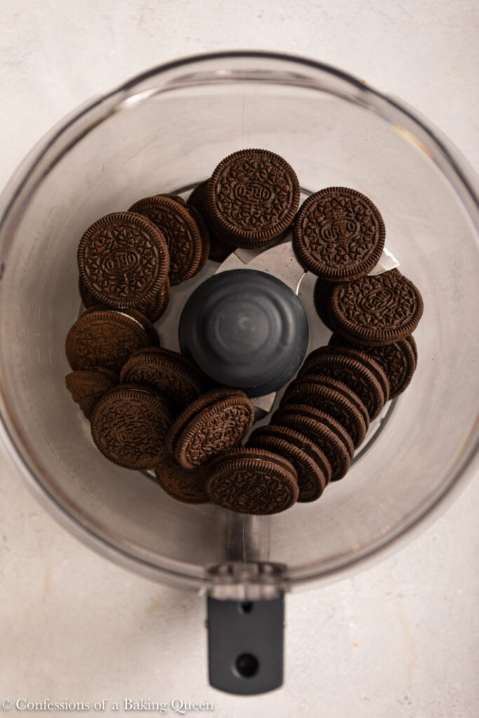 Oreos arranged in a ring inside a food processor bowl, viewed from above on a light surface—the perfect start for making rich chocolate cheesecake bars.