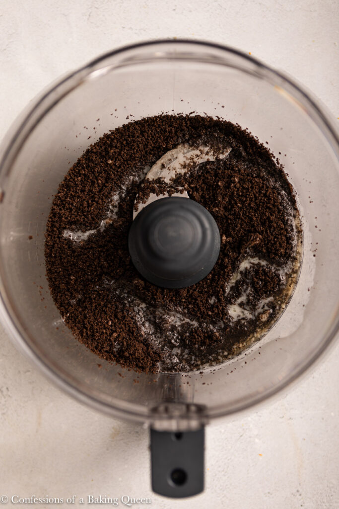 Crushed chocolate cookie crumbs and melted butter mixed together in a food processor bowl, ready to be used as a delicious crust base for chocolate cheesecake bars.