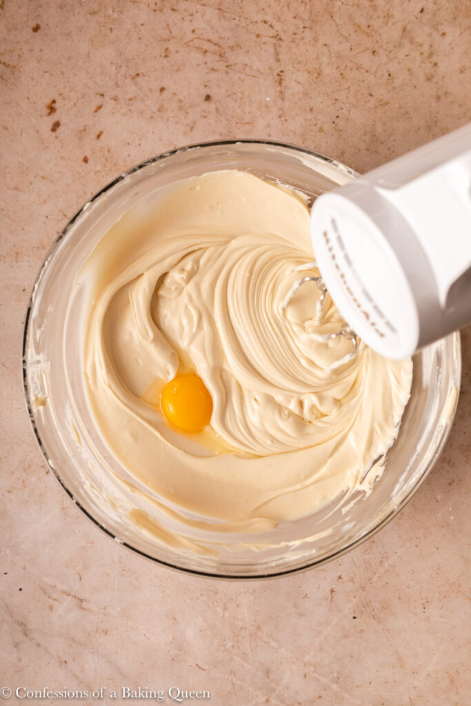 eggs added one at a time to cheesecake batter in a glass bowl with a white cordless mixer on a light brown surface