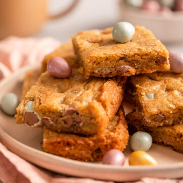 plate full of mini egg blondies on top of a light pink surface with a cup of coffee and bowl of mini eggs on alight grey surface