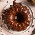 chocolate ganache frosted chocolate bundt cake served on a white plate next to a cup of coffee, knife, plates and forks