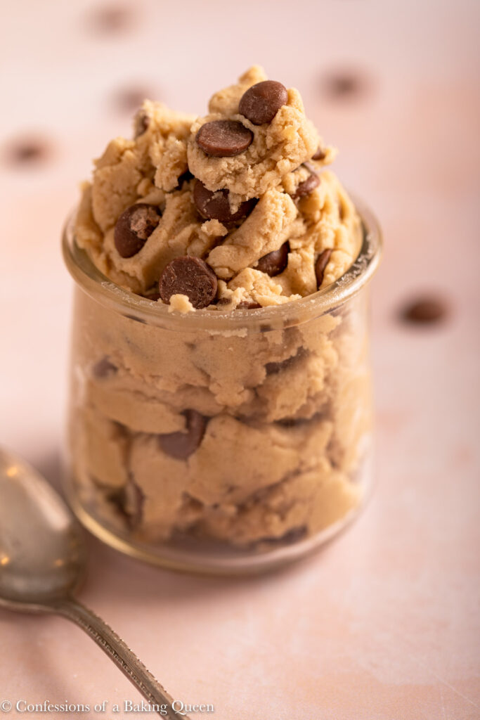 close up of edible chocolate chip cookie dough in a glass jar on a light pink surface with a spoon and extra chocolate chips