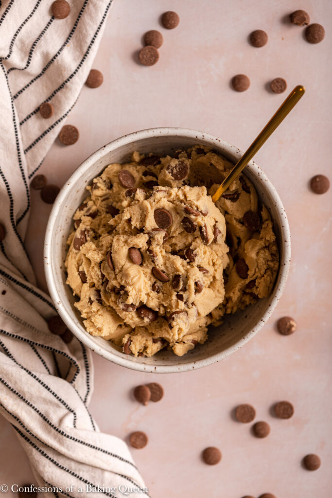 edible chocolate chip cookie dough in a ceramic bowl with a gold spoon on a light pink surface with a white stripped linen and chocolate chips all over