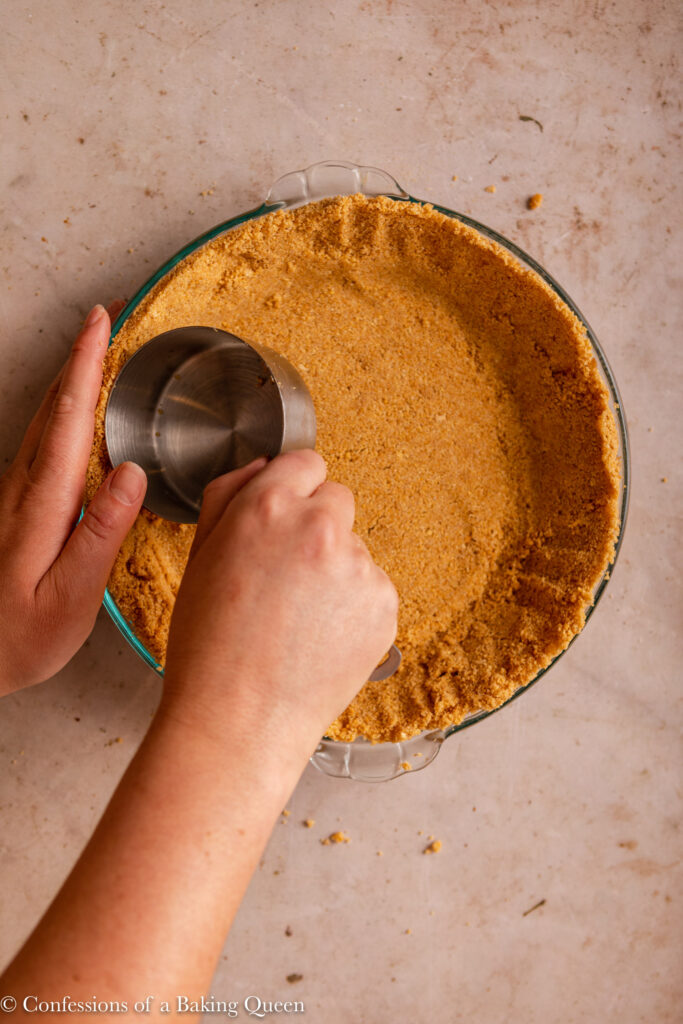 hand pressing metal cup into graham cracker crust to pack it in on a light brown surface