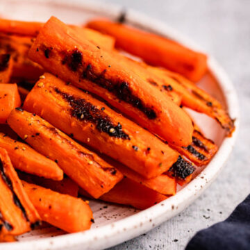 close up of roasted carrots on white and brown speckled plate on a grey surface with a navy blue linen