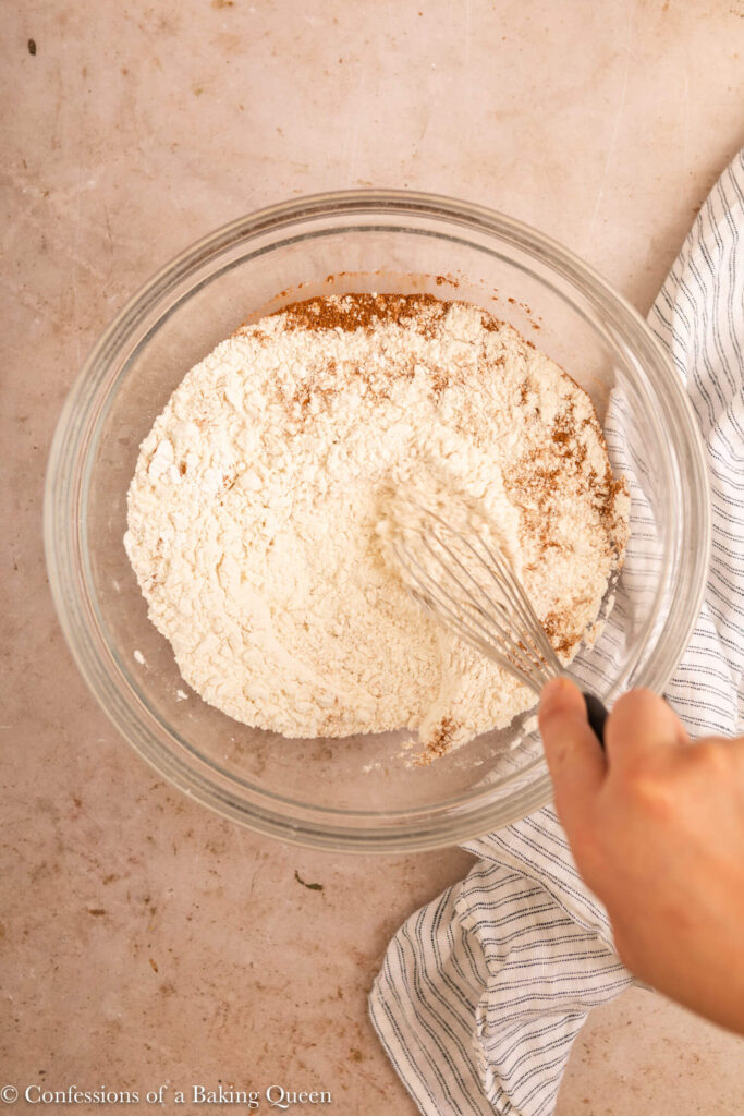 hand whisking dry ingredients together in a glass bowl on a light brown surface with a white and blue linen
