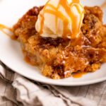 caramel apple dump cake served wtih vanilla ice cream on a white plate on top of a grey and blue linen on a marble surface