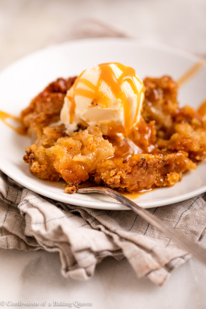fork taking a bite from a la mode caramel apple dump cake on a white plate with a fork on top of a grey and blue linen on a marble surface
