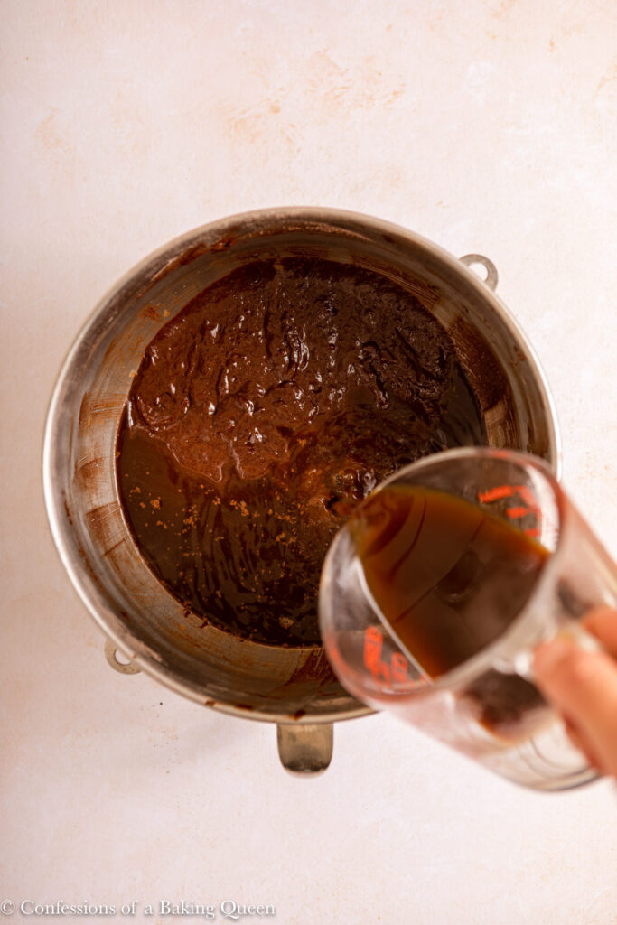 hot coffee poured into chocolate cake batter in a metal mixing bowl on a light cream surface