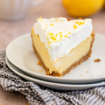 slice of lemon cream pie with whipped cream on top on a white plate on a blue linen on a light brown surface with a cup of coffee and lemons in the background