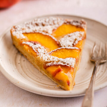 slice of powdered sugar covered peach frangipane tart on a white plate next to peaches on a light surface