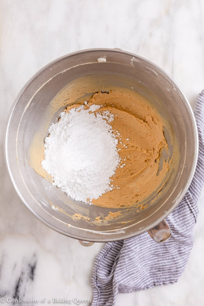 confectioners sugar added to peanut butter cream cheese mixture in a metal bowl on a white marble surface with a blue linen