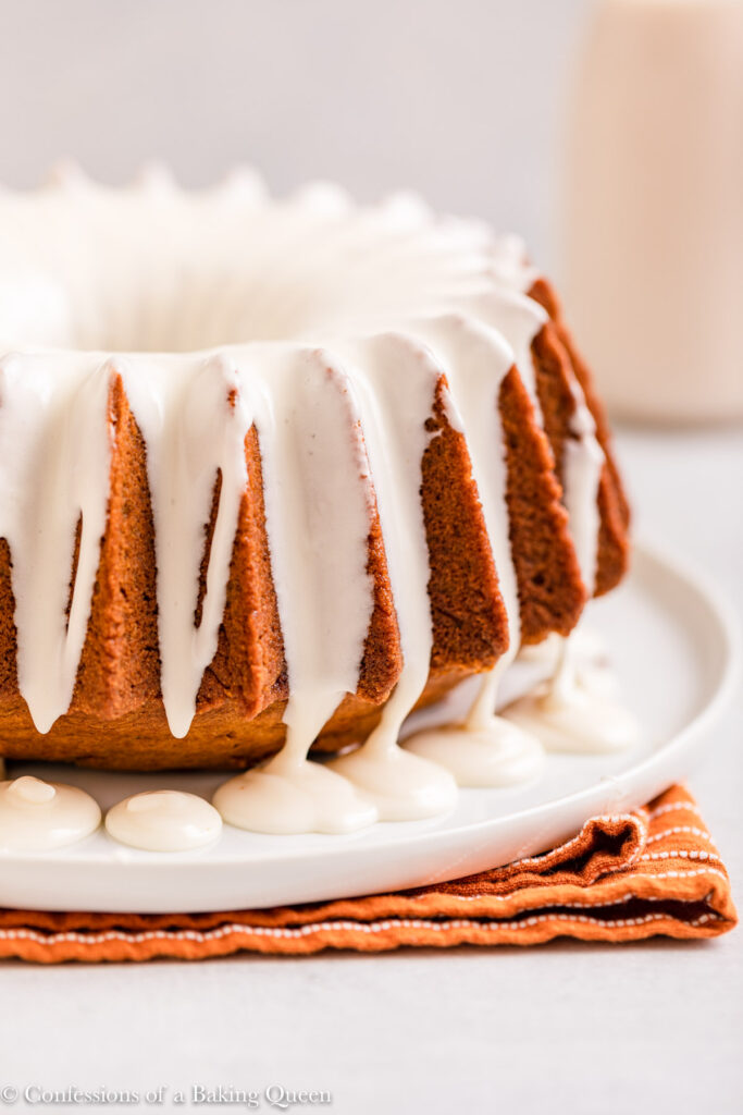 cream cheese glazed pumpkin cake on a white plate on top of an orange and white linen on a light grey surface with milk bottle in the background