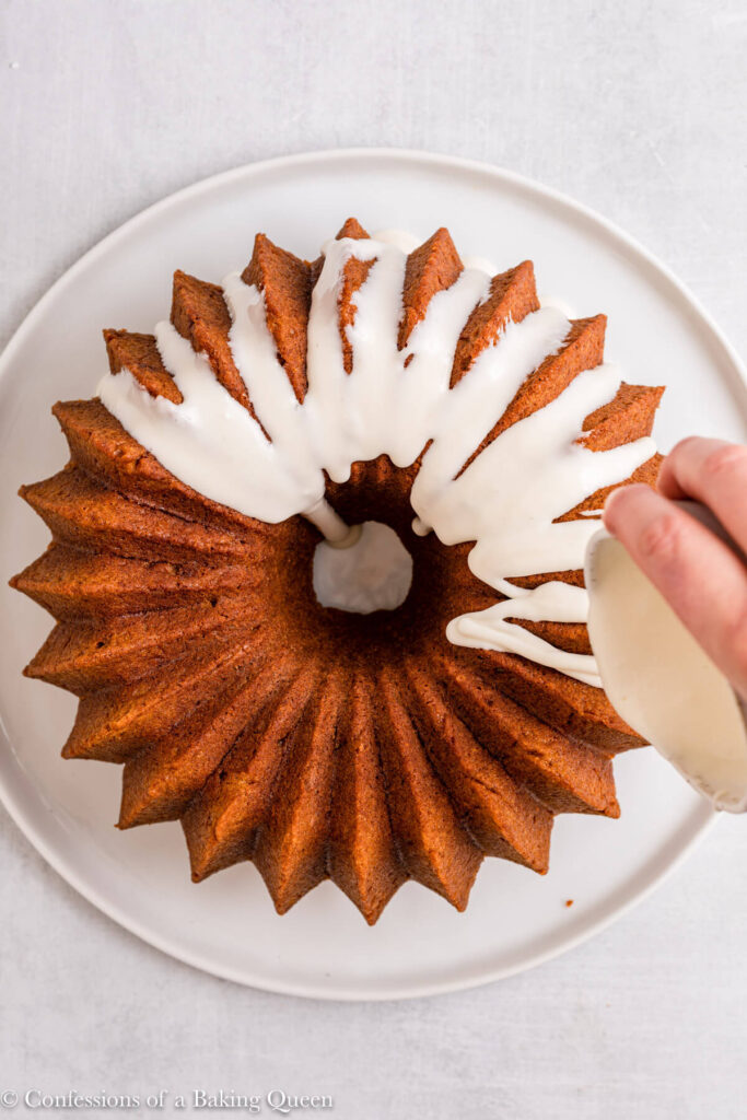 hand drizzle cream cheese glaze on pumpkin spiced bundt cake on a white plate on a light grey surface