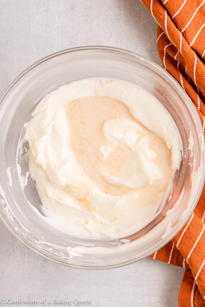 heavy cream and vanilla extract added to cream cheese frosting in a glass bowl next to an orange linen on a light grey surface
