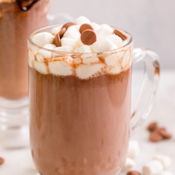 marshmallow topped hot chocolate in a large glass mug on a white marble surface next to chocolate chips, marshmallows and more hot chocolate