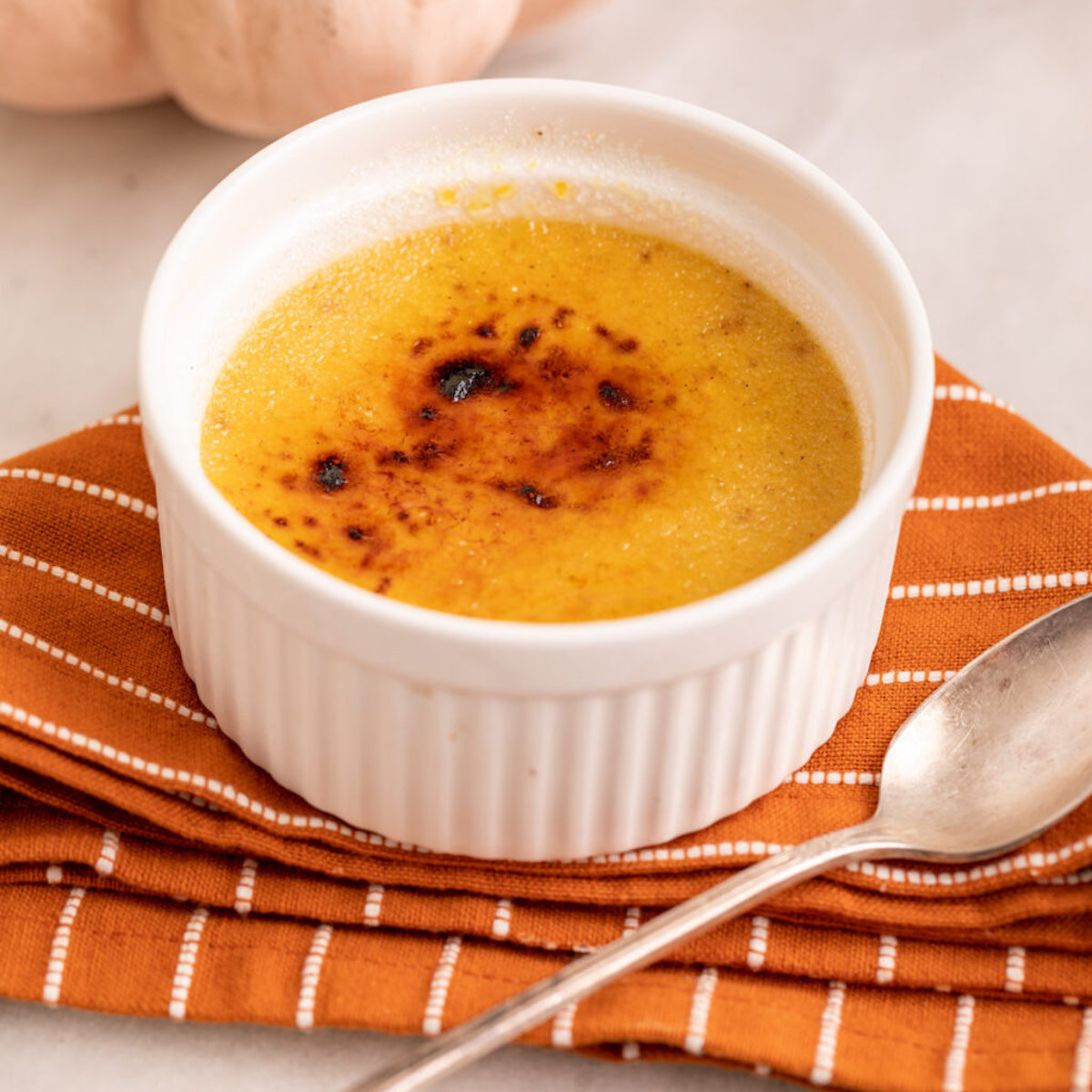 pumpkin creme brulee served next to a pumpkin on a white marble surface with an orange linen