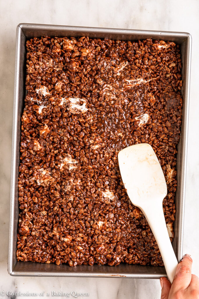 spatula pressing chocolate rice krispies into an even layer in a metal sheet pan on a white marble surface
