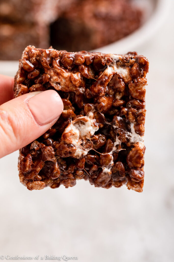 up close of hand holding a chocolate rice krispy over a white marble surface with a plate of more rice krispies in the background