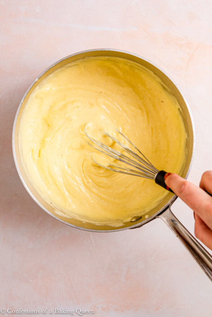 custard thickening as it cooks in a metal pot with a whisk mixing it on a light pink surface