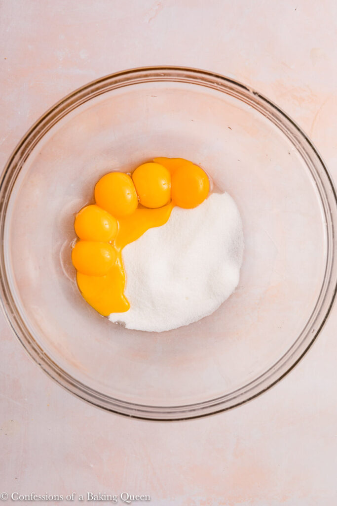 egg yolks and sugar in a glass bowl on a light pink surface