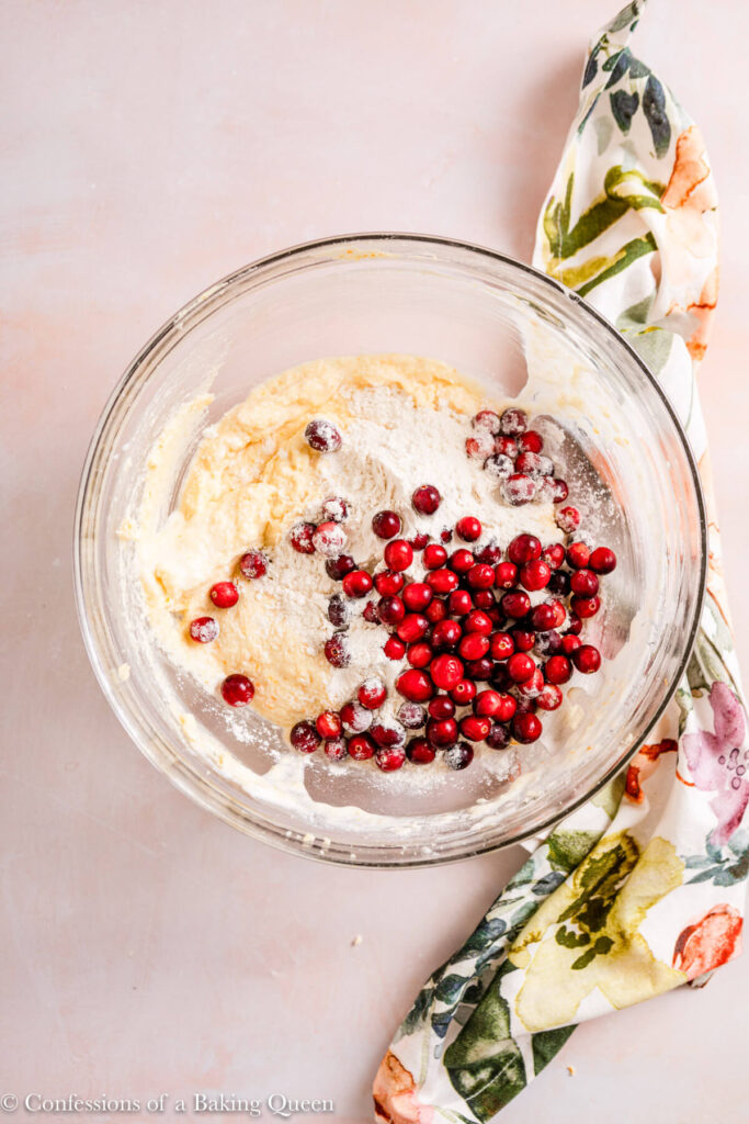 fresh cranberries and dry ingredients added to orange cake batter in a glass bowl on a light pink surface with a floral linen