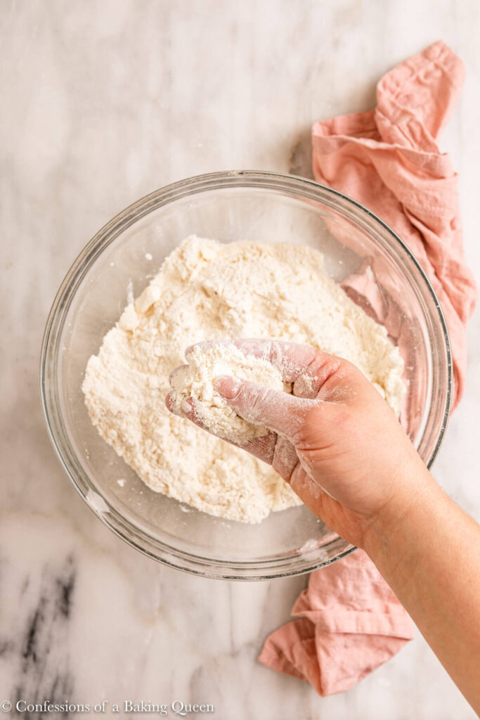 hand rubbing butter into dry ingredients in a glass bowl on a white marble surface with a pink linen