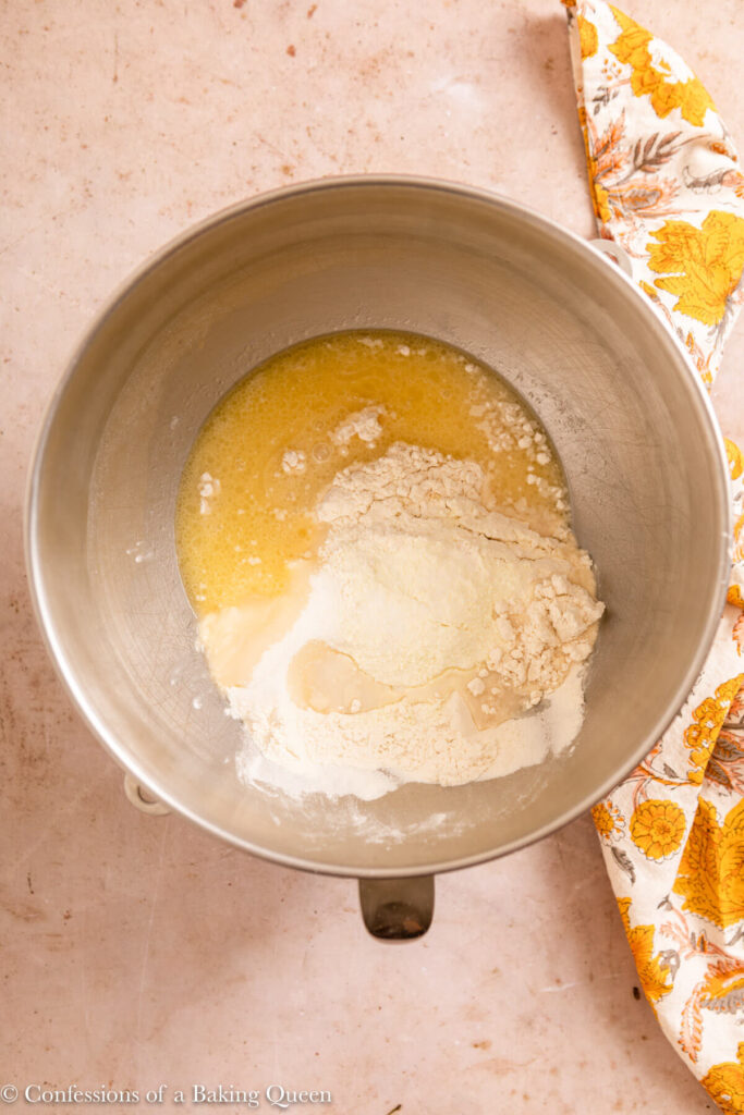 ingredients added to yeast to make sweet dough in a metal mixing bowl on a light brown surface with a yellow floral linen